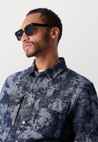 Denim shirt in blue tones with abstract pattern, featuring a button-up front, collar, and chest pockets. Black square sunglasses complete the look.