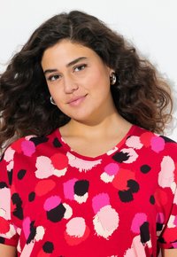 Žena s vlnitými tmavě hnědými vlasy, která nosí stříbrné kruhové náušnice a červenou košili s abstraktními kruhovými vzory v růžové, černé a bílé barvě.