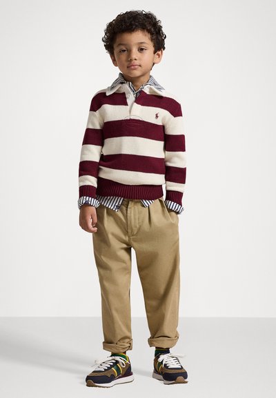 Svītrains džemperis bordo un bēšā krāsā, apvienots ar zilu un baltu svītrainu krekla apkakli. Khaki bikses un tumši zili sporta apavi noslēdz apģērbu.