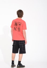 Le t-shirt corail à manches courtes présente des illustrations graphiques noires au dos. Associé à un short cargo noir et des chaussures noires.
