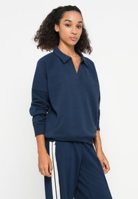 Marineblauwe pullover met een polo kraag en raglan mouwen. Zacht, textuurstof met een losse pasvorm, gecombineerd met bijpassende marineblauwe broek met witte strepen.