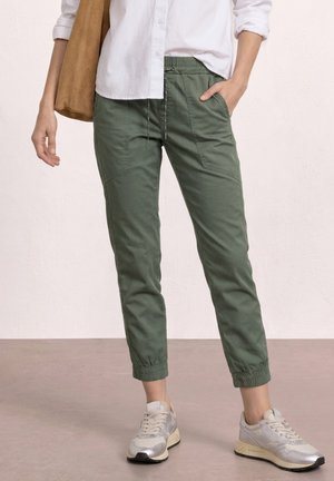 Personne portant un pantalon de jogging vert olive à cordon de serrage avec poches avant, des baskets blanches et une chemise blanche boutonnée, tenant un sac beige.