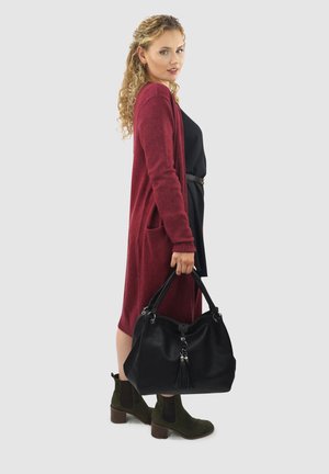 Bordeaux lange cardigan over een zwarte jurk, met zijzakken. Zwarte handtas met textuur en kwastdetail, gecombineerd met olijfgroene enkellaarsjes.