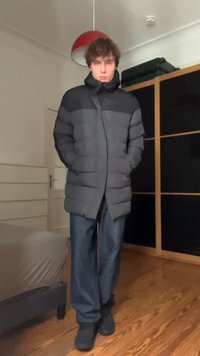 Jaunas žmogus stovi viduje, apsirengęs juodu ir pilku pūkuotu švarku, mėlynomis kelnėmis ir juodais batais, rankas įsidėjęs į švarko kišenes.