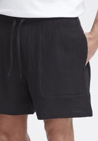 Schwarze Shorts aus strukturiertem Stoff mit Karomuster. Verfügt über einen elastischen Bund mit Kordelzug und Seitentaschen.