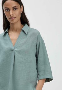 Blusa in lino verde chiaro con colletto, scollatura a V e ampie maniche a tre quarti con piega centrale e una texture sottile.