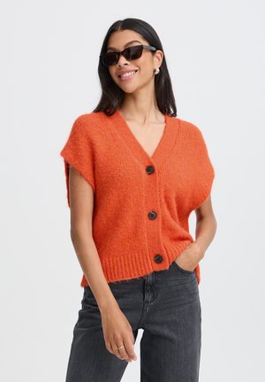 Frau mit schwarzen Sonnenbrillen, orangefarbener Strickjacke mit kurzen Ärmeln und drei Knöpfen sowie dunkler Jeans, steht mit einer Hand in der Tasche.