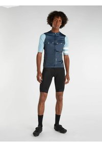 Maillot de cyclisme à manches courtes en marine avec des manches bleu clair, fermeture éclair à l'avant et motifs géométriques. Associé à un short noir rembourré et des chaussures noires.