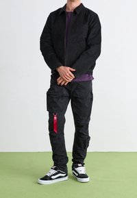 Osoba stojąca z dłońmi skrzyżowanymi, ubrana w czarne spodnie cargo z czerwonym logo, czarną kurtkę na fioletowej koszulce i czarno-białe sneakersy na zielonej podłodze.