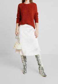 Pull en maille rouille, jupe blanche en satin et bottes hauteur genou à motif serpent. Sac transparent avec sac intérieur crème. Matériaux texturés et couleurs contrastées.