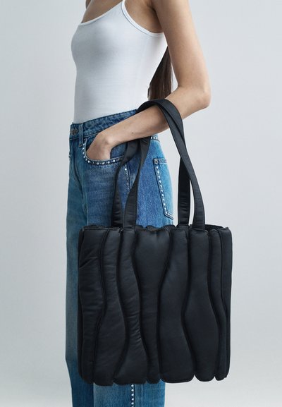 Mustat pehmeät olkalaukku, jossa on aaltoileva pinta, pehmeä nahkamateriaali ja kaksi vahvaa kantohihnaa. Yhdistetty sinisiin farkkushortseihin, joissa on koristeellisia niittejä.