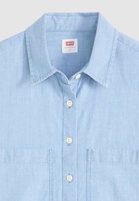 Camicia di jeans blu chiaro con colletto classico, chiusura a bottoni, due tasche sul petto e bottoni bianchi. Tessuto morbido e testurizzato.