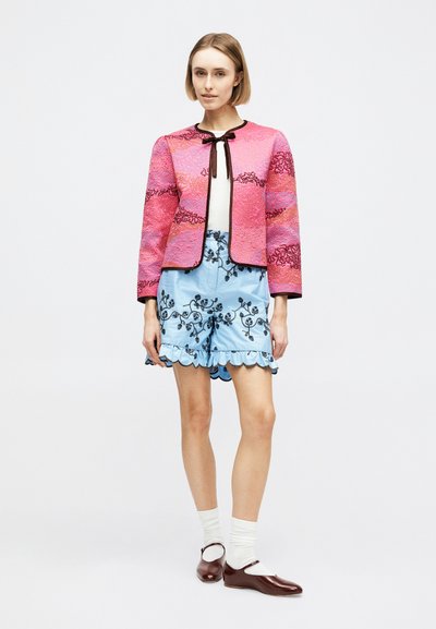 Γυναίκα που στέκεται, φοράει υφασμένη ροζ και κόκκινη ζακέτα με μαύρες λεπτομέρειες, ανοιχτό μπλε κεντημένα σορτς με κυματιστoύς γείσους, λευκές κάλτσες και καφέ παπούτσια.