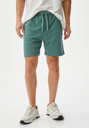 Smaragdegrønne træningsshorts lavet af blødt stof, med en elastisk talje med snor og sidelommer. Båret med lyse sneakers.