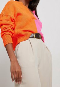Pull tricot bicolore orange et rose avec col en V, associé à un pantalon crème taille haute et une ceinture marron foncé avec une boucle dorée.