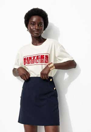 T-shirt crème avec un graphique "SISTERS" rouge, associé à une mini-jupe navy avec des accents de boutons dorés sur les côtés. Textures lisses dans l'ensemble.