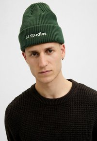 Zielona, żebrowana czapka beanie z logo "JJ Studios" w kolorze białym. Noszona z ciemnobrązowym swetrem o fakturze.