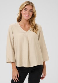 Blouse beige avec un décolleté en V, des manches trois-quarts, des détails plissés à l'avant, un tissu au toucher doux et une coupe décontractée.
