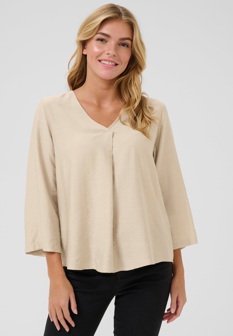 Blouse beige avec un décolleté en V, des manches trois-quarts, des détails plissés à l'avant, un tissu au toucher doux et une coupe décontractée.