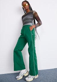 Πράσινα, ελαφριά παντελόνια με ελαστική μέση και λευκές ρίγες στο πλάι, με λογότυπο Nike, συνδυασμένα με ένα μακρυμάνικο τοπ με σχέδια και αθλητικά παπούτσια.