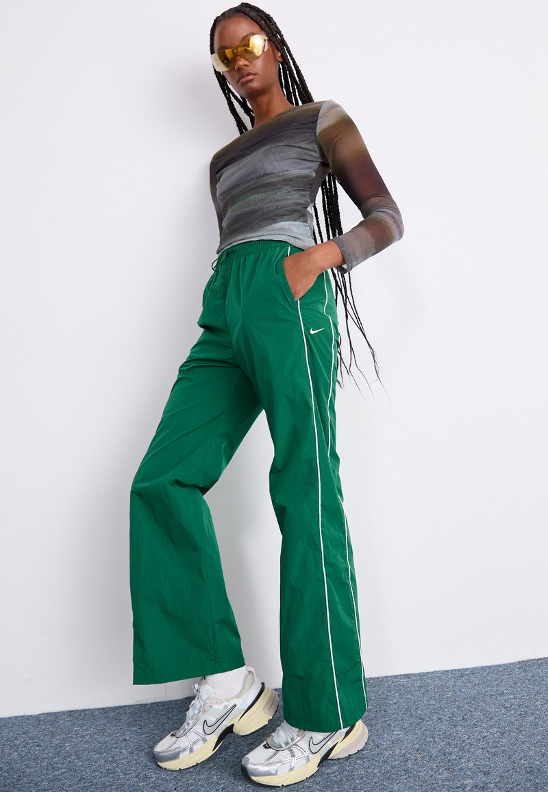 Groene, lichte, elastische broek met witte zijstrepen en het Nike-logo, gecombineerd met een gestreept longsleeve shirt en sneakers.