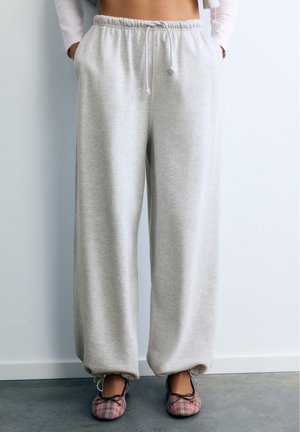Personne portant un pantalon de survêtement gris clair à cordon et des ballerines roses à carreaux, debout contre un mur blanc.