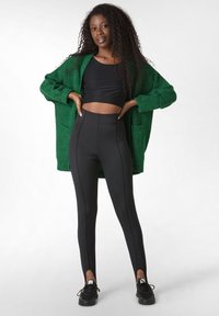 Zelený pletený kardigan přes černý crop top, doplněný o černé legíny s vysokým pasem s bočními rozparky a hladkou texturou. Černé tenisky.