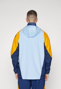 Šarena jakna na vjetar s svijetloplavim tijelom, žutim i tamnoplave naglašava, podesivim kapuljačom, elastičnim manžetama i opuštenim krojem.