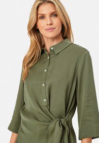 Olivenfarbene Bluse mit weicher Textur, Knopfleiste an der Vorderseite und Dreiviertelärmeln. Sie verfügt über einen gebundenen Akzent in der Taille, der für Form sorgt.