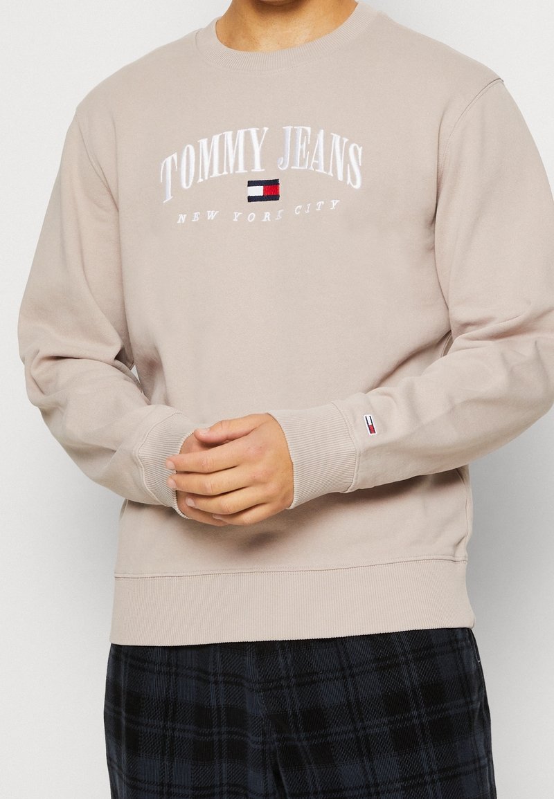 Beżowa bluza z okrągłym dekoltem, z haftowanym białym napisem „TOMMY JEANS NEW YORK CITY” oraz małym logo na mankiecie.