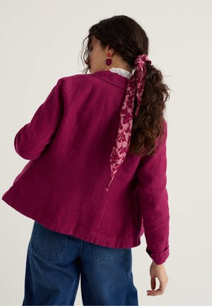 Femeie cu păr creț prins cu un fular roz cu flori, purtând o jachetă roz închis, cercei mov și blugi albaștri, cu spatele către cameră.