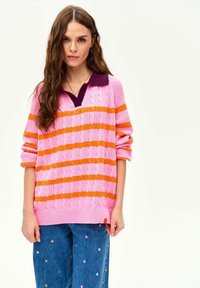 Młoda kobieta o długich włosach, ubrana w różowo-pomarańczowy sweter w paski z ciemnym kołnierzem oraz niebieskie jeansy z małymi kwiatowymi wzorami.