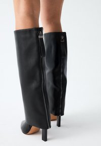 Botas de cuero negro hasta la rodilla con una textura lisa, diseño con cortes en la parte frontal, acabado brillante y cremalleras en la parte posterior. Los tacones cuadrados acentúan la silueta.
