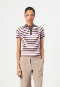 Ung kvinna med flätat hår, iförd en kortärmad polo i rosa, vitt och brunt ränder samt beige byxor, står mot en vit bakgrund.