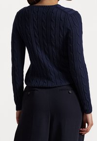 Marinblå kabelstickad tröja med långa ärmar, texturerat mönster, ribbad nederkant och stickad detalj längs axlarna. Kombinerad med mörka byxor.