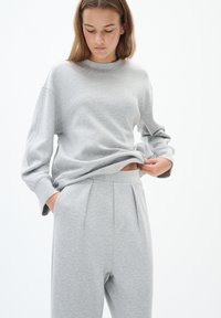 Grijze sweatshirt met lange mouwen en een ronde halslijn, gecombineerd met bijpassende hoge, geplooide broeken. De stof lijkt zacht en gestructureerd.