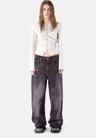 Junge Frau mit welligem Haar, die ein gerafftes weißes Oberteil mit Knöpfen, übergroße dunkelviolette Jeans und schwarze flache Schuhe trägt und gerade steht.