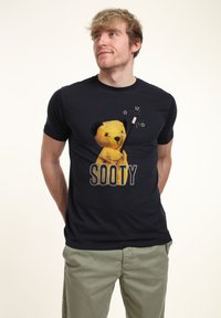 Svart T-shirt med en grafik av Sooty björnen i gult som håller en trollstav, med "SOOTY" i fet, prickig bokstavering nedanför.