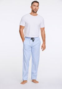 Valkoinen lyhythihainen t-paita yhdistettynä sinisiin raidallisiin pyjamahousuihin, joissa on joustava vyötärö ja musta naru. Alasti jaloillaan, seisoo vaalealla taustalla.