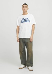 Hvid t-shirt med "JACK & JONES" i navy grafik. Bred, falmet denimbukser, casual sneakers. Neutral baggrund, minimale accessories.
