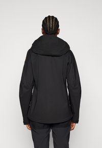 Veste noire imperméable avec capuche, présentant une texture lisse et une coupe ajustée. Des poches latérales zippées et des poignets réglables sont visibles.