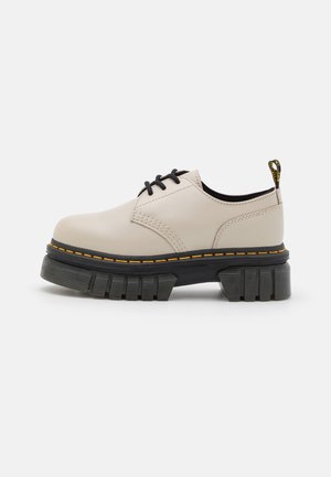 Chaussure beige en cuir à lacets avec semelle épaisse noire et couture jaune, vue de côté sur fond blanc.