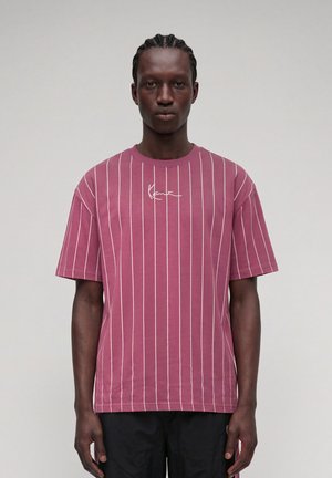 Mand med fletninger bærer en sart lilla T-shirt med tynde hvide lodrette striber og et lille hvidt logo, stående mod en ensartet lys baggrund.