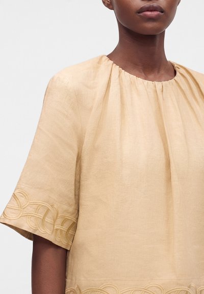 Femme portant un top en lin beige clair à manches courtes avec des motifs circulaires brodés près des bords des manches et de l’ourlet.
