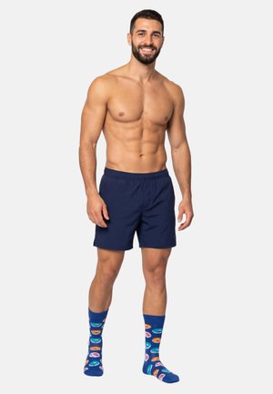 Man zonder shirt met baard, gekleed in marineblauwe korte broek en kleurrijke sokken met donutpatronen, glimlachend, staand tegen een effen lichte achtergrond.