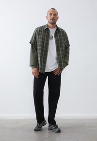 Short-sleeve plaid shirt in green and blue over a white t-shirt, paired with black pants and gray sneakers. Features a relaxed fit and layered look.