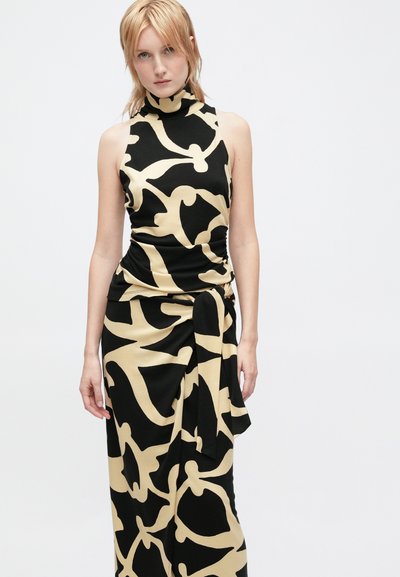 Jovem mulher vestindo um vestido sem mangas com um alto decote e um grande padrão floral abstrato em bege e preto, de pé contra um fundo simples.