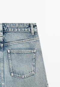 Achteraanzicht van lichtblauwe spijkerbroeken met een enkele gestikte zak en riemlussen op de tailleband tegen een witte achtergrond.