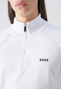 Femme portant un pull blanc à fermeture éclair avec une tirette noire et le logo "BOSS" sur la poitrine gauche, sur un fond clair uni.