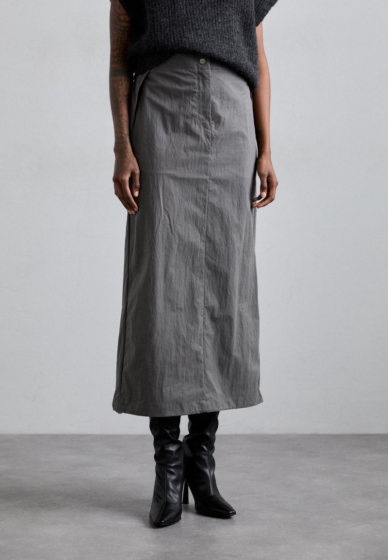 Jupe grise jusqu'aux genoux en tissu texturé, dotée d'une fermeture à boutons et d'une silhouette droite, associée à des bottes à talons noires.